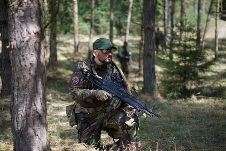 A solider wearing a camo outfit in the woods while holding an automatic gun on his knee staring at a distance next to a tree.